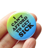 A round 1.25" pinback button with a green to blue ombre that reads I CANT AFFORD TO GET SICK in thick black text. Button is held in a hand.