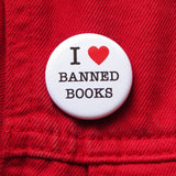 A round white pinback button that reads I LOVE BANNED BOOKS. Love represented by a red heart. Button is on a red denim jacket.