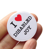A round white 1.25" pinback button that reads I LOVE DISABLED JOY in a black serif font, the love being indicated by a red heart. Badge is in a hand.