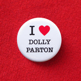 A round, white pinback button that reads I love Dolly Parton. Love being indicated with a red heart. Button is pinned to a red sweatshirt.