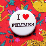 A round white pinback button that reads I LOVE FEMMES in a black serif font, the love being indicated by a red heart. Button is on red silk shirt patterned with leopards and florals.