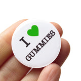 A round white pinback button that reads I LOVE GUMMIES, has a green heart representing love, and the rest of the phrase is in a classic serif font. The button is held in a hand.