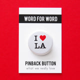 A round white pinback button that reads I LOVE LA. Love represented by a red heart. Button is on a Word For Word branded backing card that says Pinback Button.