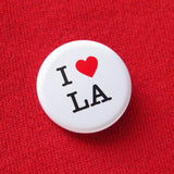 A round white pinback button that reads I LOVE LA. Love represented by a red heart. Pinned to a red sweatshirt.
