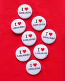 Eight round white 1.25" pinback button that reads I LOVE LIBRARIES, love being represented by a red heart. Buttons are pinned to a red sweatshirt.