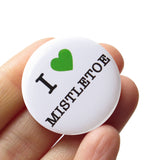 Round, white pinback button that says I LOVE MISTLETOE in classic black text with "love" being represented by a green heart. Button is held in a hand. 