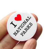 A round white pinback button that reads I LOVE NATIONAL PARKS. Love represented by a red heart. Button is held in a hand.