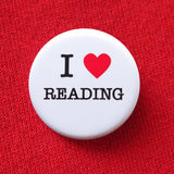 A round white pinback button that reads I LOVE READING, love being represented by a red heart. Button is held in a hand.