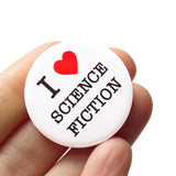 Round white pinback button that reads I LOVE SCIENCE FICTION. "Love" is represented by a red heart. Button is held in a hand.