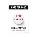 A round white button that reads I LOVE SNACKS. Love being represented by a red heart. Button is pinned to a Word For Word branded backing card.