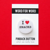 A round white button that reads I LOVE SNACKS. Love being represented by a red heart. Button is pinned to a Word For Word branded backing card.