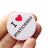 A round white pinback button that reads I LOVE STATIONERY, with love being indicated by a red heart. Badge is held in a hand