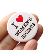 Round white pinback button that says I love women artists with a red heart. Button held in a hand.