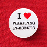 Round, white pinback button that says I LOVE WRAPPING PRESENTS in classic black text with "love" being represented by a red heart. Button is pinned to a red sweatshirt