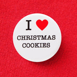 Round, white pinback button that says I LOVE CHRISTMAS COOKIES in classic black text with "love" being represented by a red heart. Button is pinned to a red sweatshirt