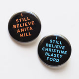 Two round black pinback buttons - one  reads I STILL BELIEVE ANITA HILL in peach text, the other in I STILL BELEIVE CHRISTINE BLASEY FORD.