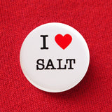 A round white button that reads I LOVE SALT. Love being represented by a red heart. Button is pinned to a red sweatshirt