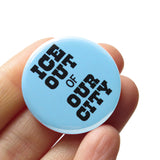 Round light blue pinback button that reads: ICE OUT of OUR CITY in chunky black text. Badge is held in a hand.