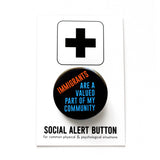 Round black pinback button that reads: IMMIGRANTS ARE A VALUED PART OF MY COMMUNITY in red-orange and blue text. The button is pinned to a Social Alert Button backing card.