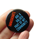 Round black pinback button that reads: IMMIGRANTS ARE A VALUED PART OF MY COMMUNITY in red-orange and blue text. Button is held in a hand.
