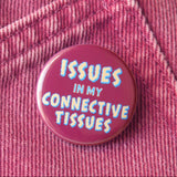 A burgundy pinback button that reads ISSUES IN MY CONNECTIVE TISSUES in a jagged cream and light blue text. Button is pinned to dark rose denim.