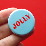 Round, pale blue-green pinback button that reads JOLLY in bright red text. Button is held in a hand in front of a background. 