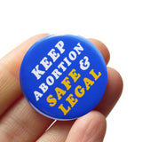 A round bright blue button that reads Keep Abortion Safe & Legal in retro yellow & white text. Button is held in a hand.