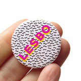 A round 1.25" white button with tiny Memphis style squiggles as a background pattern. Reading LESBO in bold pink & yellow text. Badge is held in a hand.