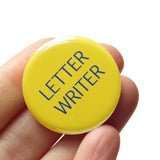 Round, light yellow button that reads LETTER WRITER in two lines in thin blue text. Badge is held in a hand.