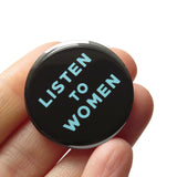 A round black pinback button that reads LISTEN TO WOMEN in light aqua blue text. Button is held in a hand.