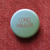 A round pale blue green pinback button that reads LONG HAULER in dark orange thin text. Button is pinned to rust orange denim. For those suffering with long covid.