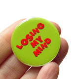 Round, lime green 1.25" pinback button off balance red text reading LOSING MY MIND. Badge is held in a hand.
