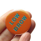 Round milk chocolate brown pinback button with aqua teal text that reads LOW BROW. Badge is held in a hand.