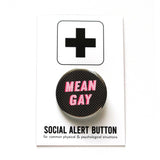 Round black pinback button with tiny white polkadots background and reads MEAN GAY in light pink with a dark pink drop shadow. On a white backing card, black plus sign at the top. Below reads: Social Alert Button, with tiny text beneath it which says, for common physical & psychological situations