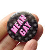 Round black pinback button with tiny white polkadots background and reads MEAN GAY in light pink with a dark pink drop shadow. Button is held in a hand.