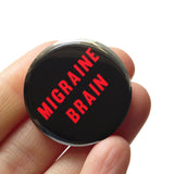 A round black pinback button that reads MIGRAINE BRAIN in red text on two lines. Button is held in a hand.