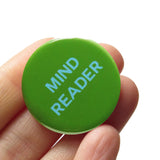 Round mossy lime green 1.25" pinback button that reads MIND READER in light blue text. Badge is held in a hand.