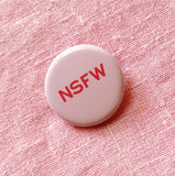 A round light pink pinback button with dark pink text that reads NSFW stands for Not Safe For Work. Button is on a pale pink linen dress.