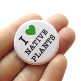 A white hand holds a  white pinback button that reads I LOVE NATIVE PLANTS, with love being represented by a green heart. 