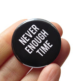 A round black pinback button that reads NEVER ENOUGH TIME in white text. Button is held in a hand.