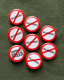 A bunch of round white 1.25" pinback buttons with red outlines and slashes indicating being against the word on the button face. Most of the buttons read NO BILLIONAIRES, and one reads NO CORPORATE GREED. Buttons are pinned to an army green fabric.