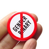 A round white button that reads No GENDER BINARY in black text with a red circle and slash to indicate being against endless greed. Badge is held in a hand.