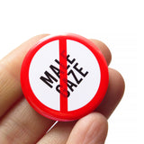 A round white button that reads NO MALE GAZE in black text with a red circle and slash to indicate being against the male gaze, or END THE MALE GAZE. Button is held in a hand.