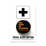 Round black pinback button that reads: NO ONE IS ILLEGAL ON STOLEN LAND in mustard yellow and red-orange text. The button is pinned to a Social Alert Button backing card.