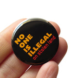 Round black pinback button that reads: NO ONE IS ILLEGAL ON STOLEN LAND in mustard yellow and red-orange text. Button is held in a hand.