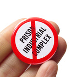 A round white button that reads NO PRISON INDUSTRIAL COMPLEX in black text with a red circle and slash to indicate being against the prison industrial complex. Badge is held in a hand.