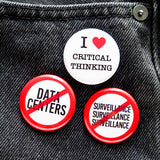 Three red, white & black round pinback buttons pinned to a black denim pocket. Buttons include: A round white pinback button with a red outline circle-and-slash graphic over the letters 'DATA CENTERS' in black text, indicating the wearer is against Data Centers & AI artificial intelligence.  A round white pinback button with a red outline circle-and-slash graphic over the letters 'SURVEILLANCE" three times in a row' in black text. And a round white button that reads I LOVE CRITICAL THINKING
