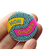 Round pinback button reading NON-BINARY.  The buttons has a  teal background and tiny black dots, a yellow ribbon comes in on the left reading NON, and a pink ribbon on the right comes in reading BINARY. The button is held in a hand.