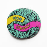 Multicolor circular stickers held at the left corner with a white thumb and forefinger. Sticker backgrounder is teal with tiny black dots. A ribbon of yellow comes from the left side and reads NON and a ribbon of pink comes in on the right side and reads BINARY. Sticker has a retro eighties Memphis design style. 