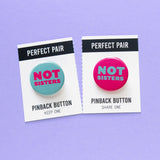 Two pinback buttons on a backing card with a center perforation to tear & share with a friend. Button on the left reads NOT SISTE with a teal background and magenta pink text. Right sided button reads NOT SISTERS in lteal with a magenta pink background.